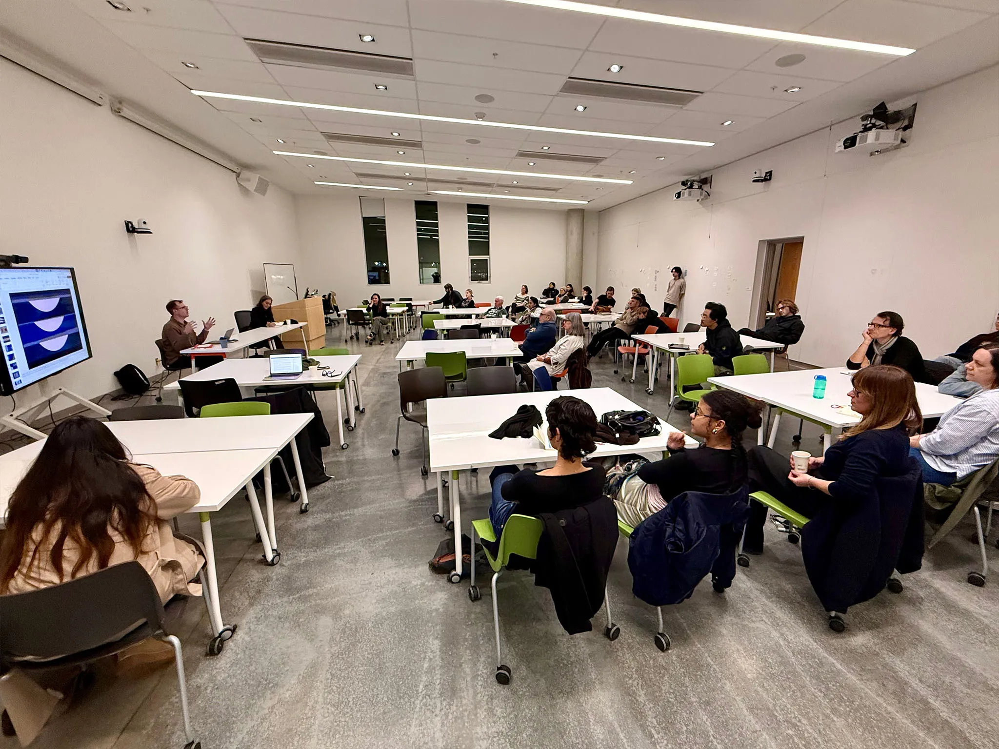 Two people speaking to a room of people in a white and grey room. Behind them is a large screen displaying images from the Ripples series.