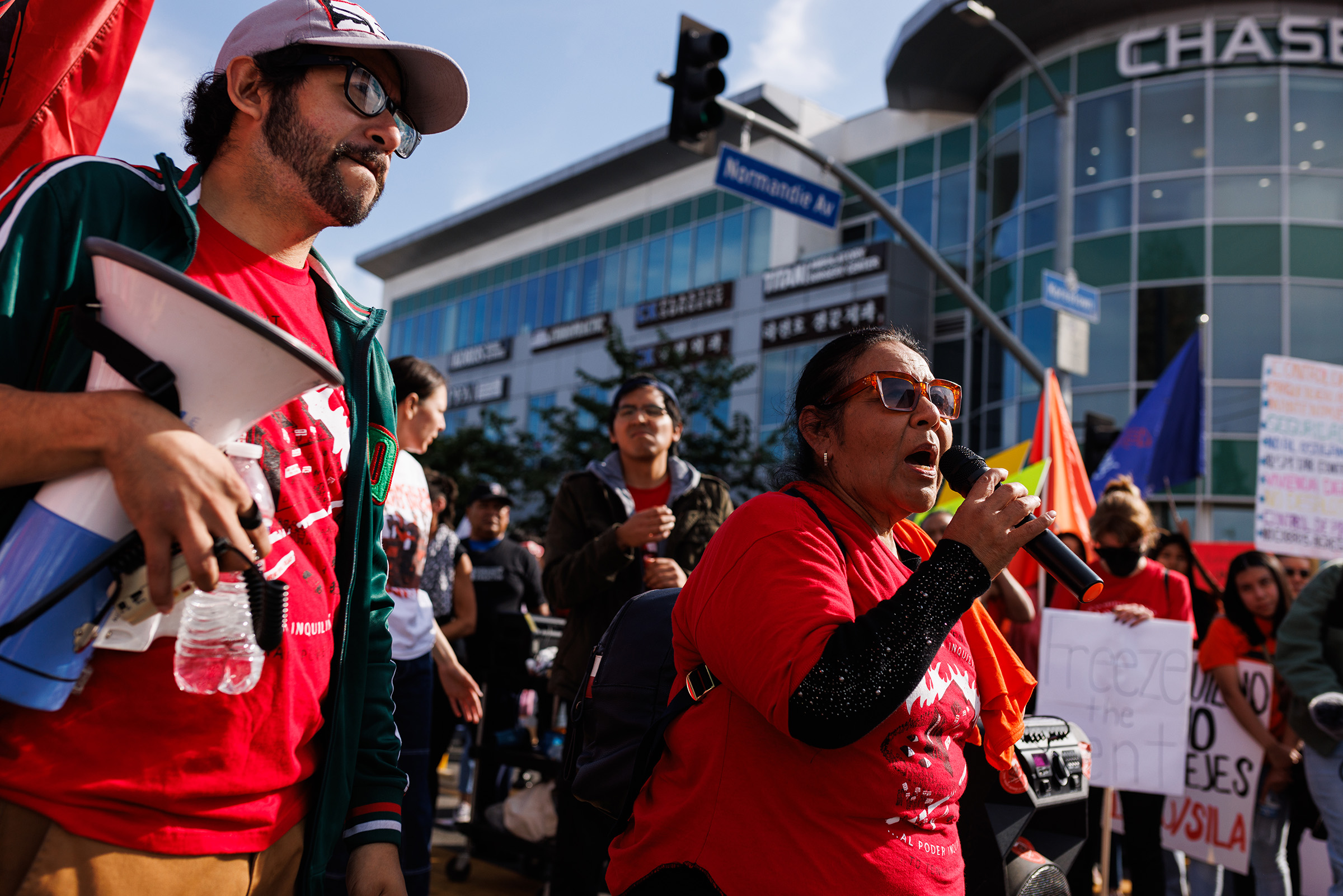 A woman in a red shirt speaking into a microphone to a group of people who are protesting rent hikes.